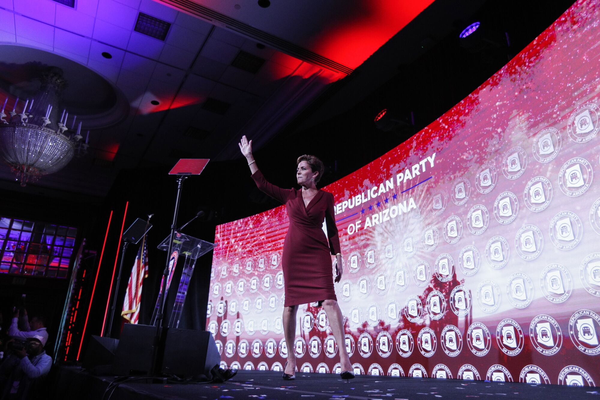 A woman waving as she walks across a stage with a red and white backdrop reading 