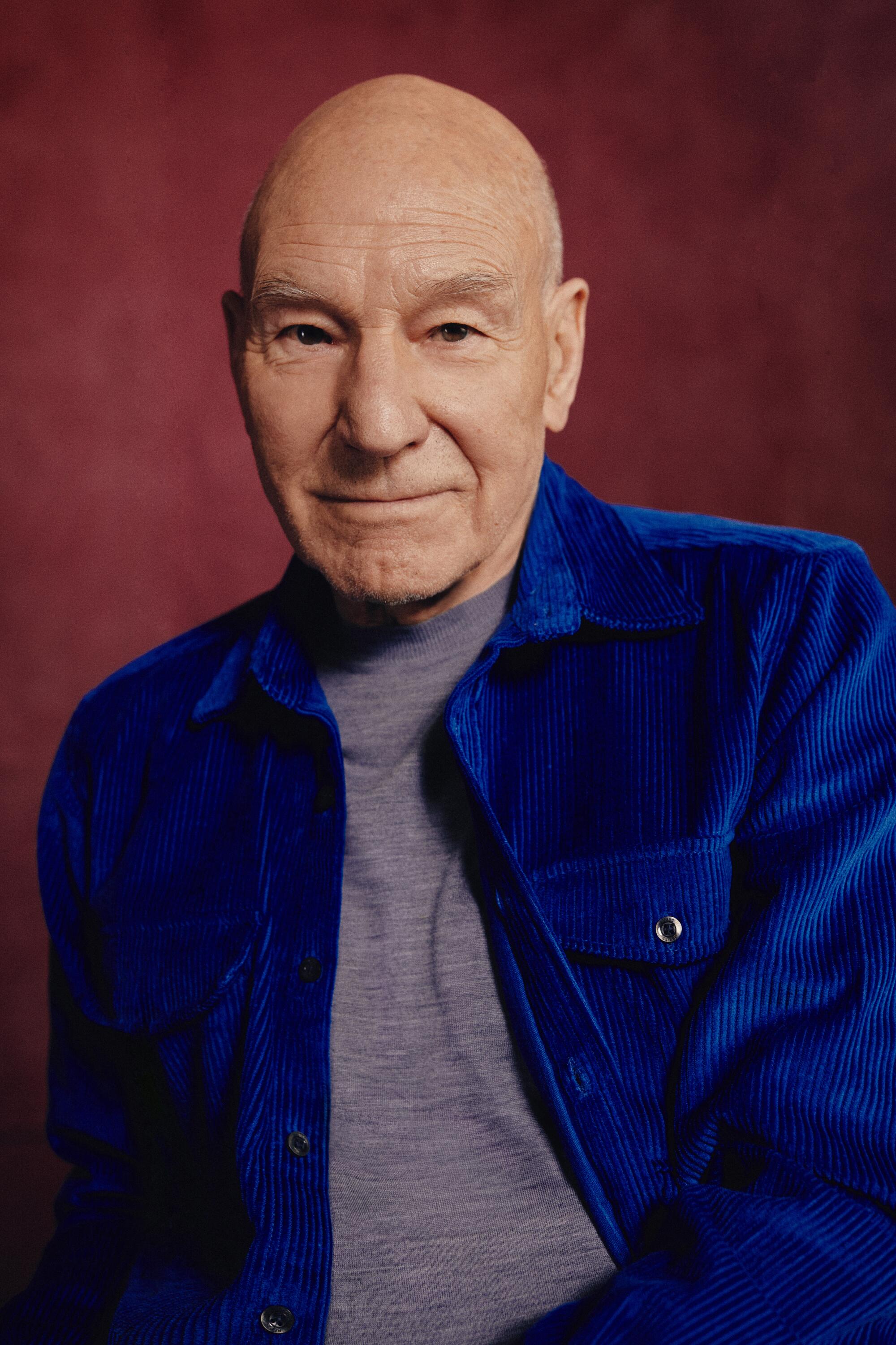 Actor Patrick Stewart in a dark blue jacket and gray T-shirt before a burgundy backdrop for a portrait.