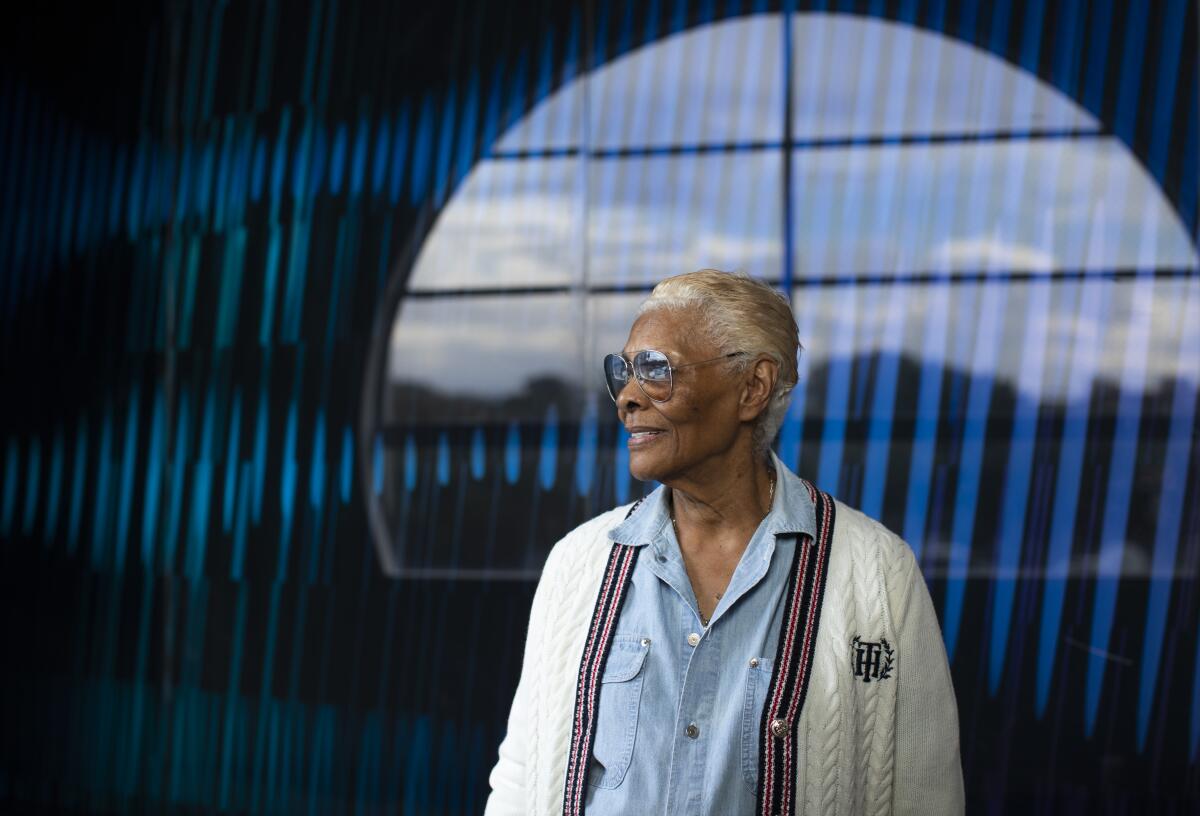 Dionne Warwick wears sunglasses and a white cardigan in front of a blue wall.