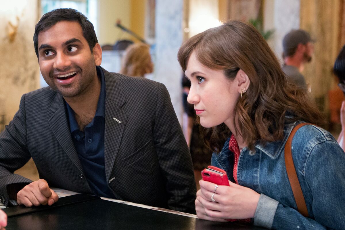 A smiling man and a woman holding a cellphone stand in front of a desk.