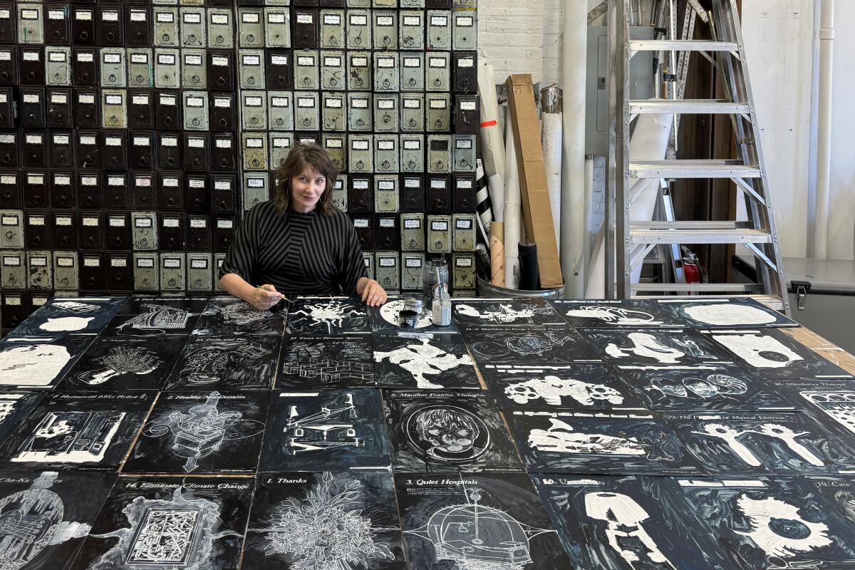 A woman sits at a vast table with black and white illustrations spread out before her. 