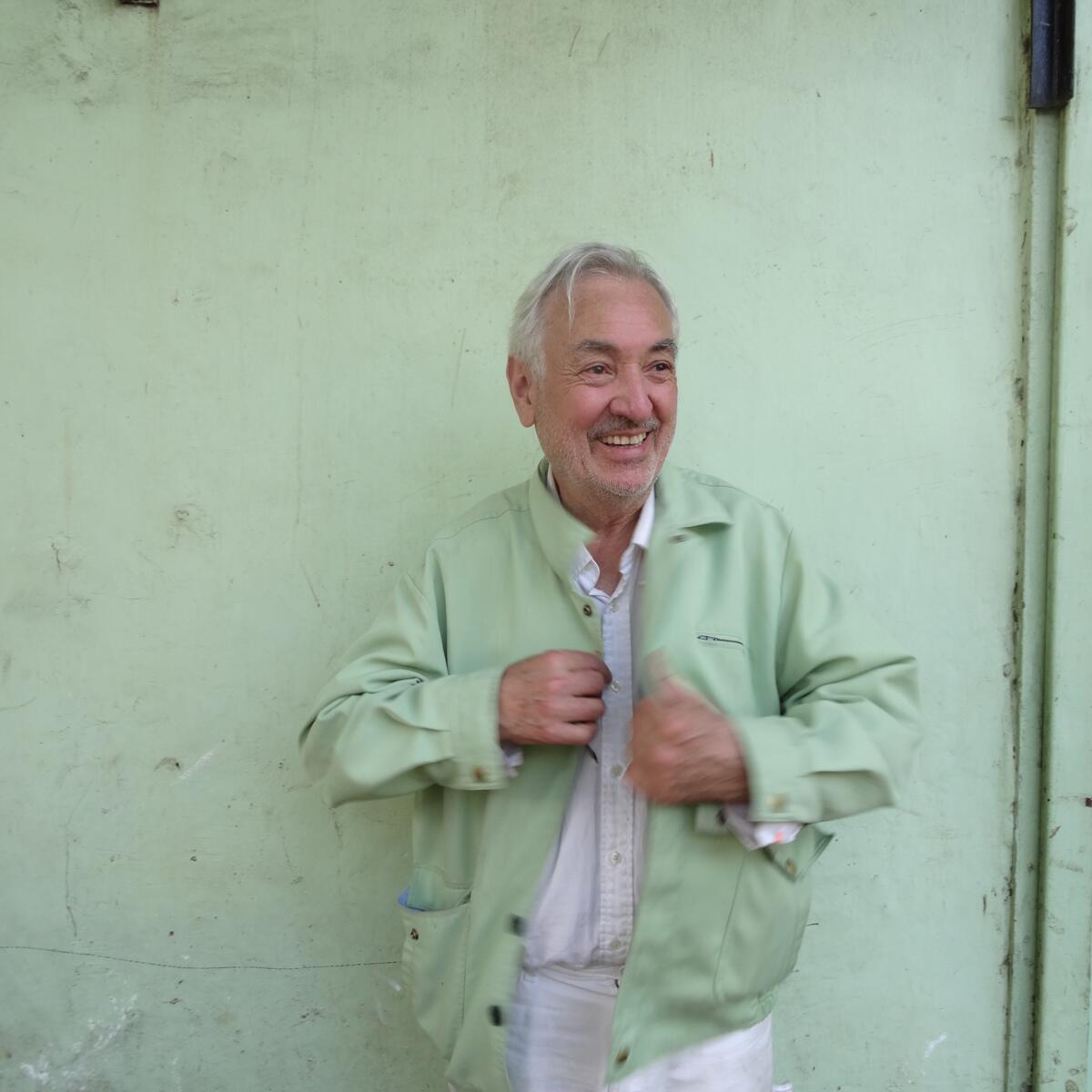 A man with gray hair wearing a mint green jacket smiles before a mint green wall