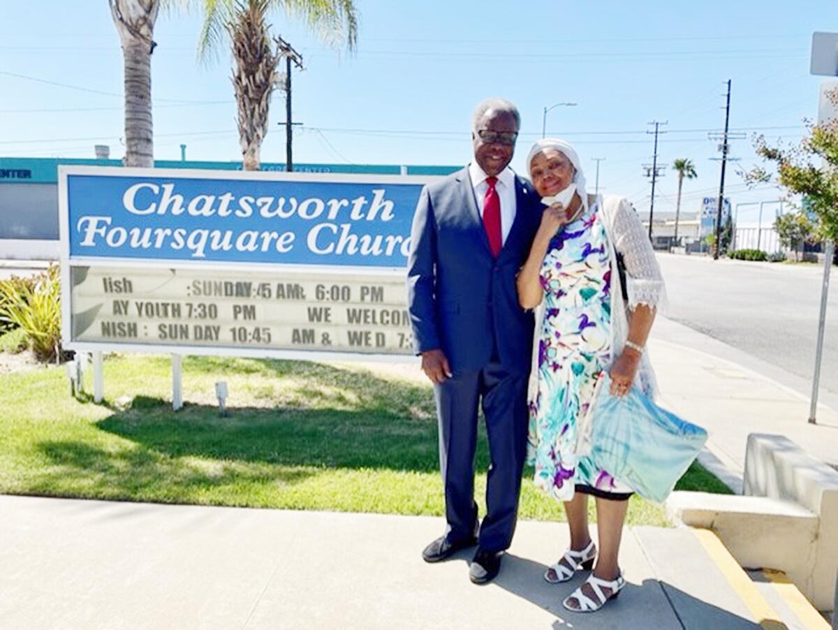 A man in a suit and a woman in a flowered dress stand outside alongside a church sign.