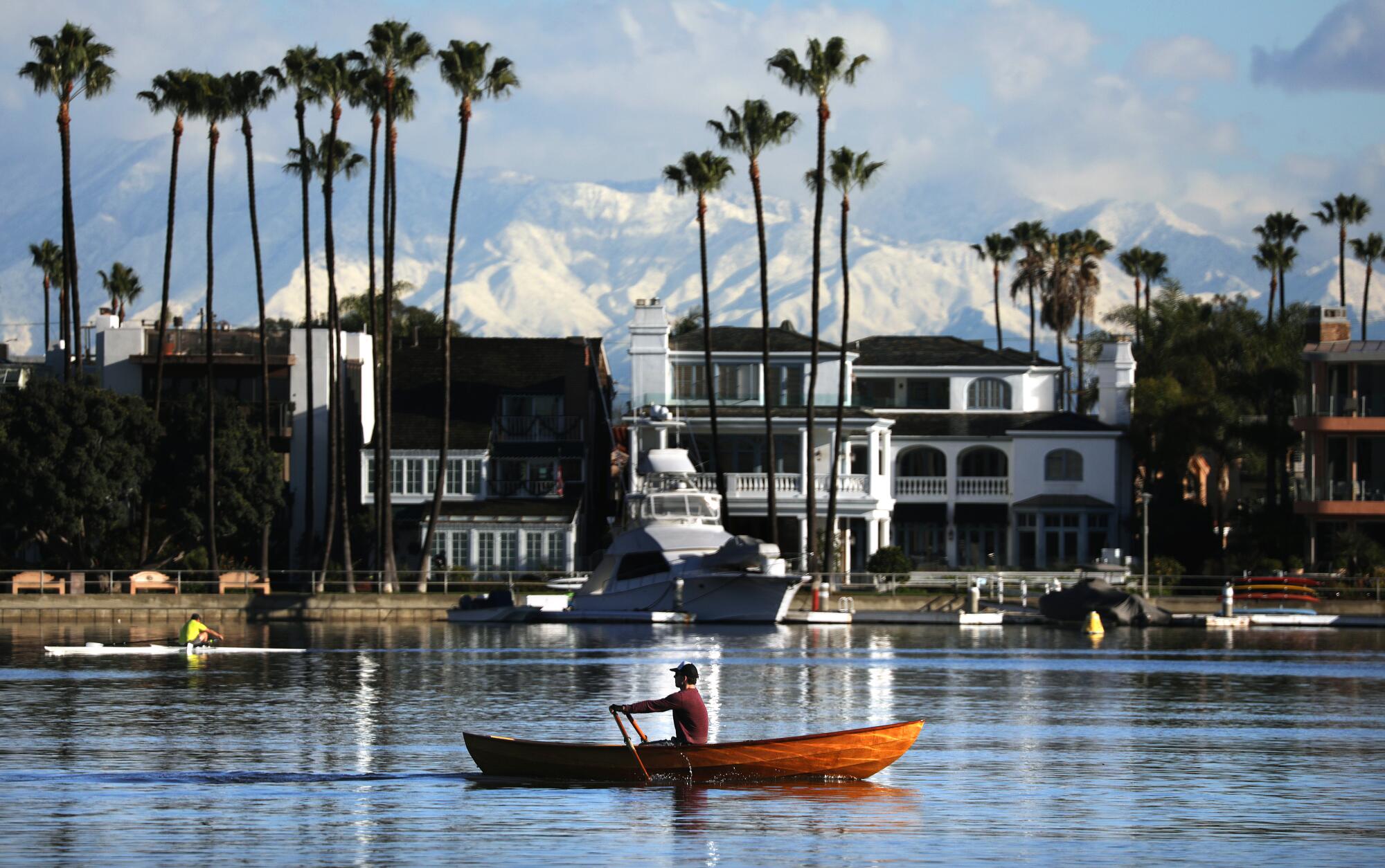 Snow-covered mountains are seen from the Peninsula in Long Beach, where a man rows a boat on the water in front of buildings.