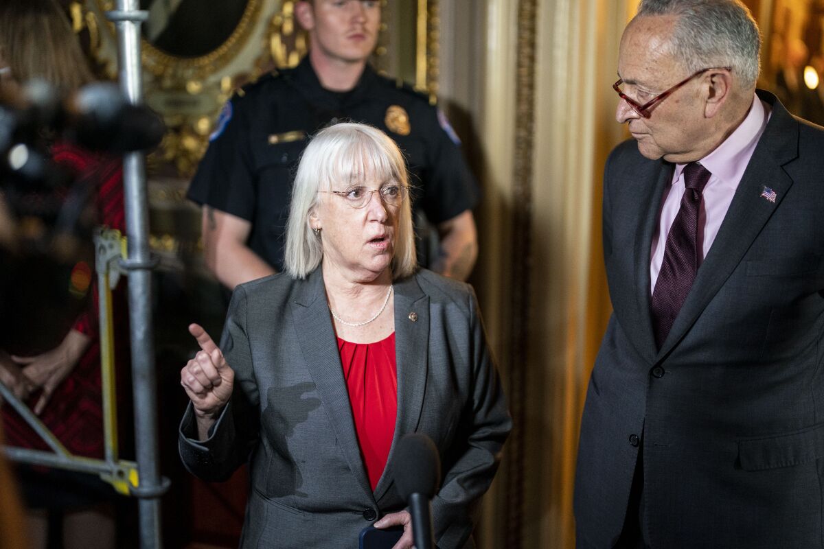 A woman in a red top and gray jacket speaks as a bespectacled man looks on