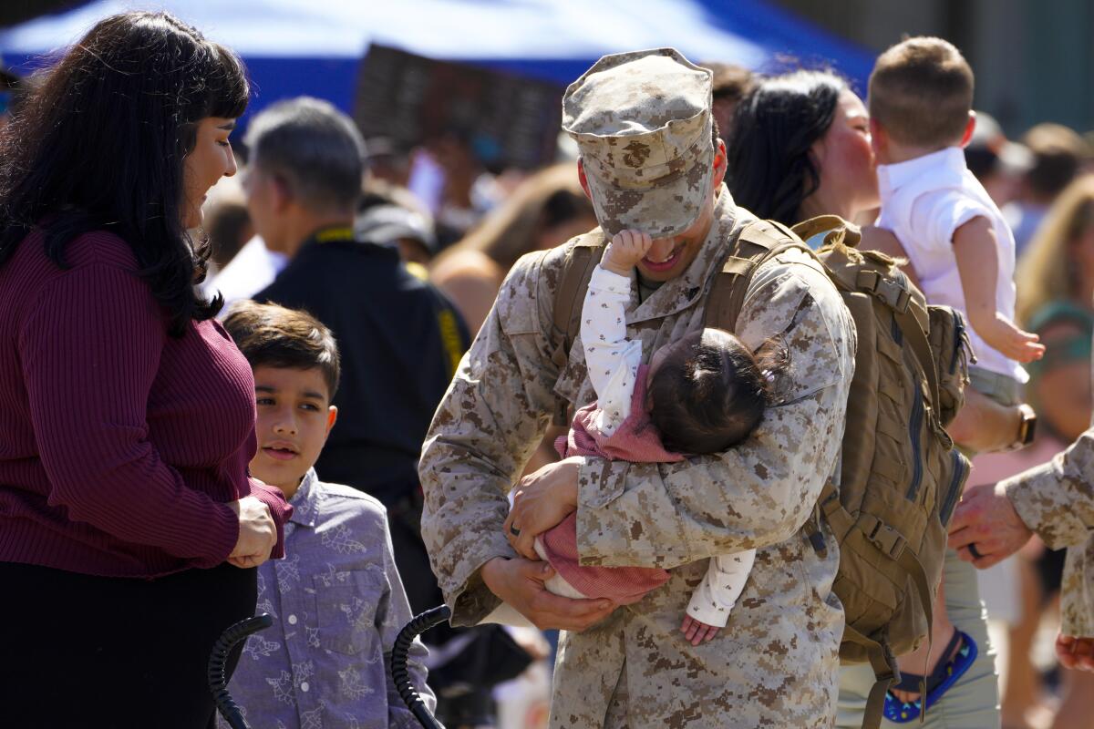 A man in camouflage uniform holds a baby as a woman and boy look on