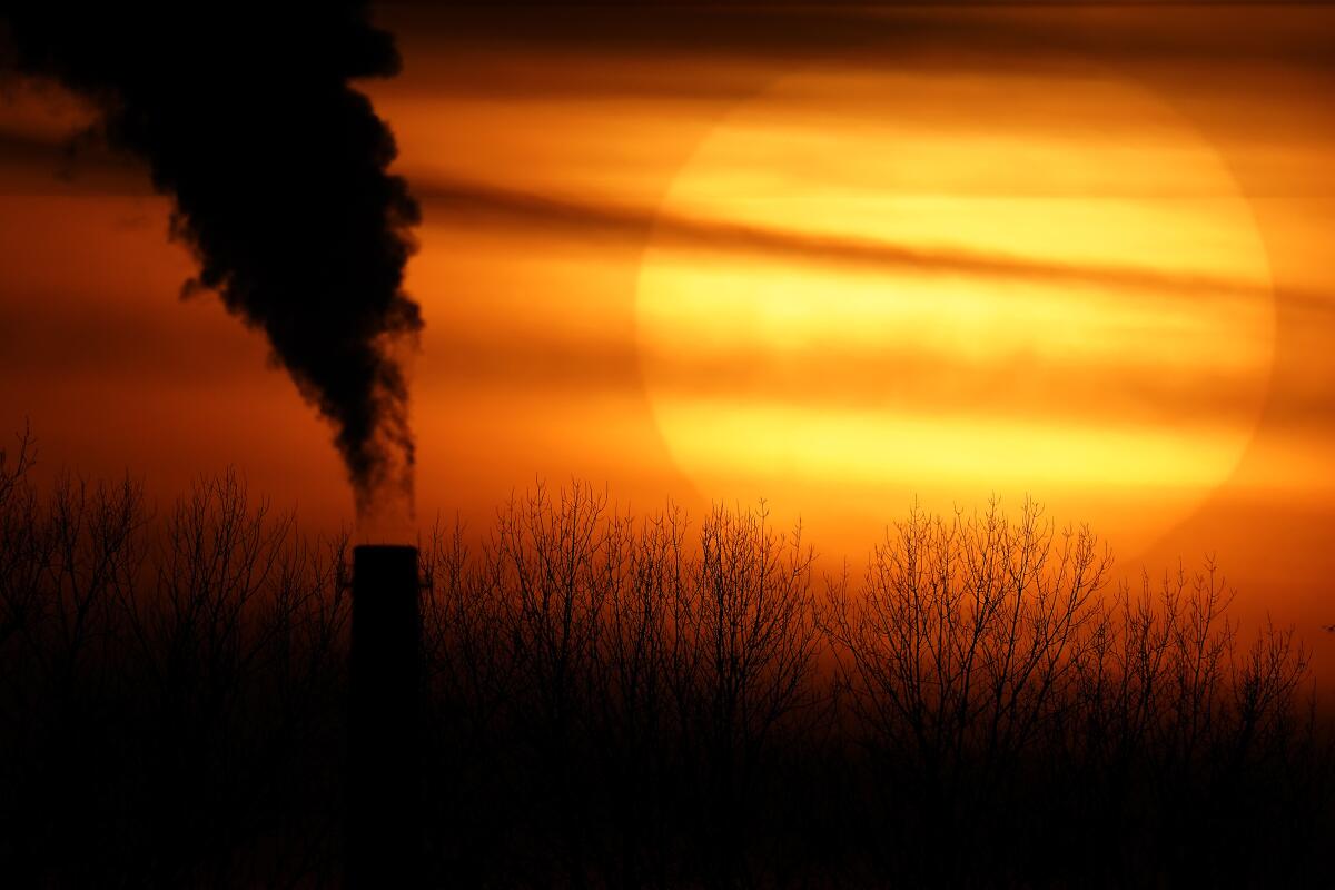 Black smoke rises from a smokestack as the sun sets in an orange-red sky.