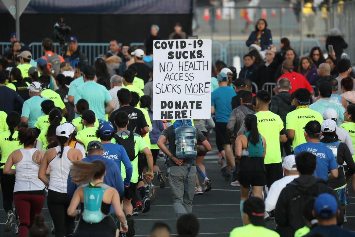 Among runners, a man carries on his back a sign that reads "COVID-19 sucks...." 