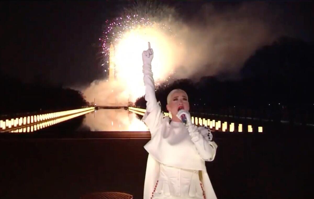 A woman pointing a finger into the air and singing into a microphone in front of fireworks and the Washington Monument.