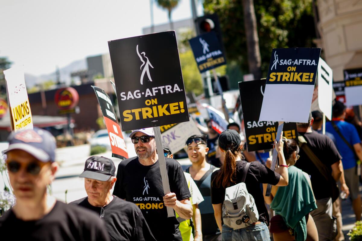 A dozen people march in line in the sunshine. Many have with signs that read SAG-AFTRA on Strike! 