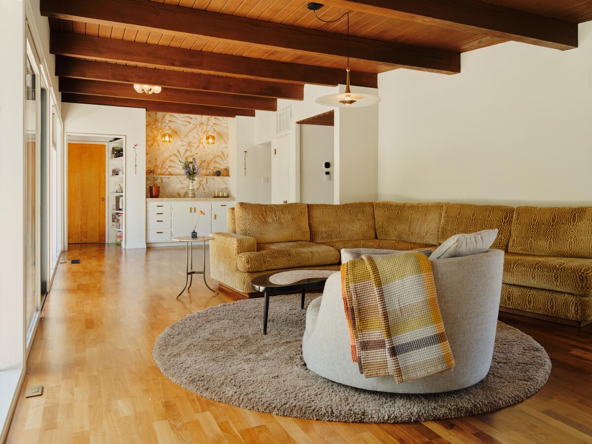A living room with wood ceiling beams and a wood floor, curved sofa and round rug, with the kitchen beyond