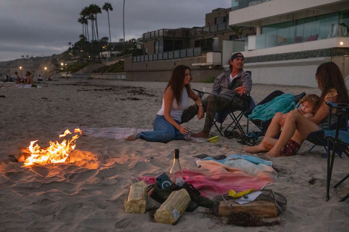 Four people lounge in chairs on the beach at twilight next to a small bonfire and a bundle of logs