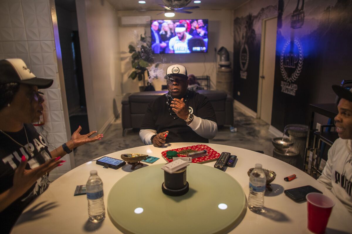 Three people sit around a round table while one of them smokes cannabis. A basketball game is playing on a TV in the background