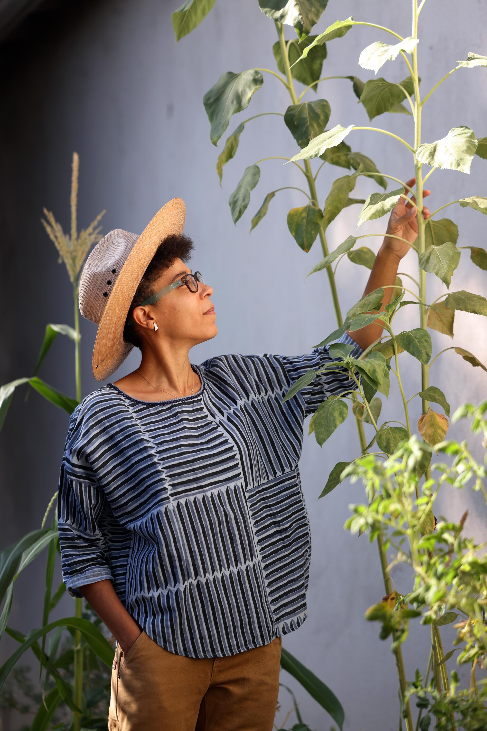 A woman wearing a straw hat stands next to tall plants.