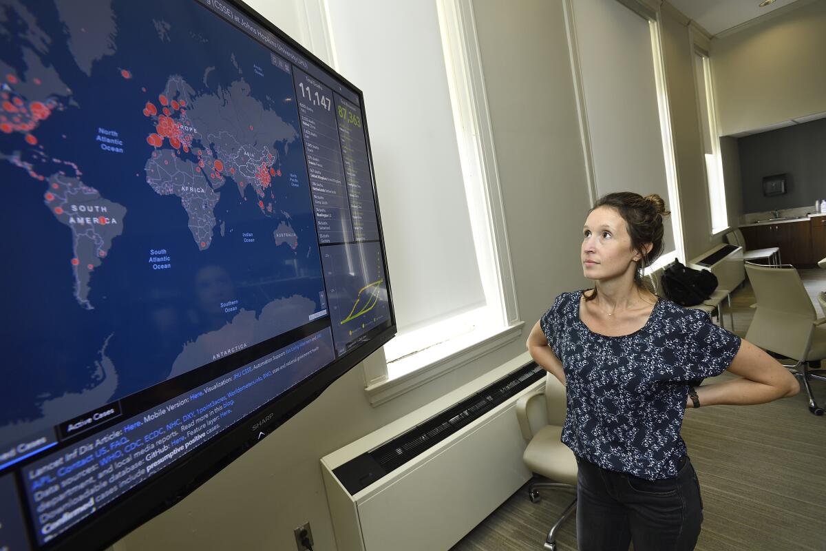 A woman with her hands on her hips looking up at a large monitor showing a world map with red spots in various locations.