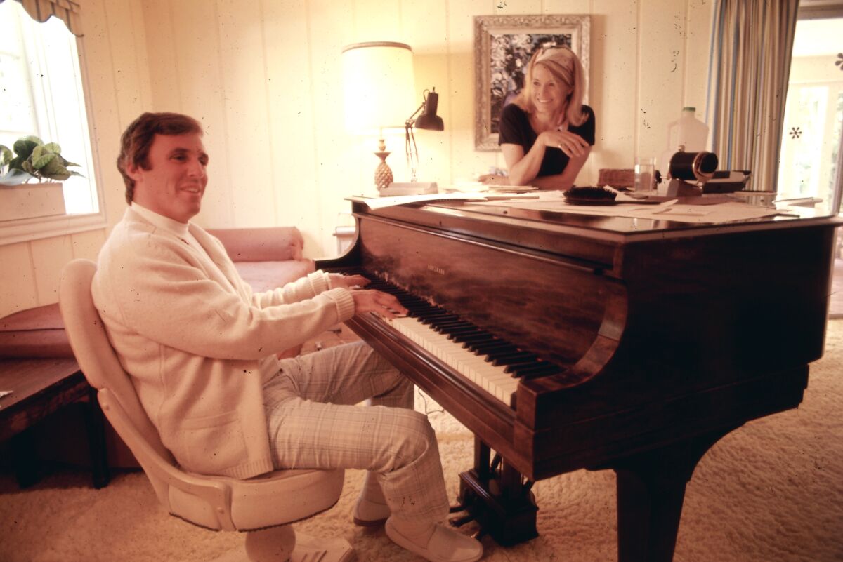 A man in a white suit plays the piano at his home while a blond woman leans on the piano watching him. 