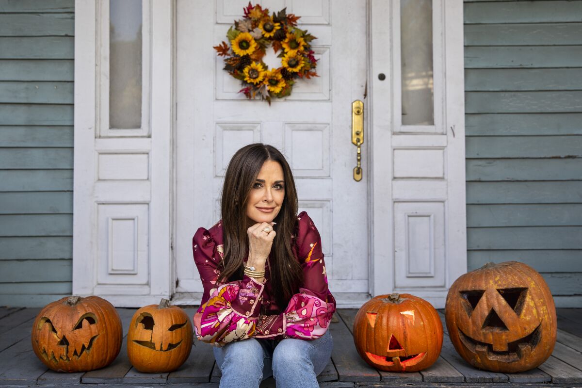 A woman with long dark hair sits on a porch decorated with carved pumpkins and a fall wreath.