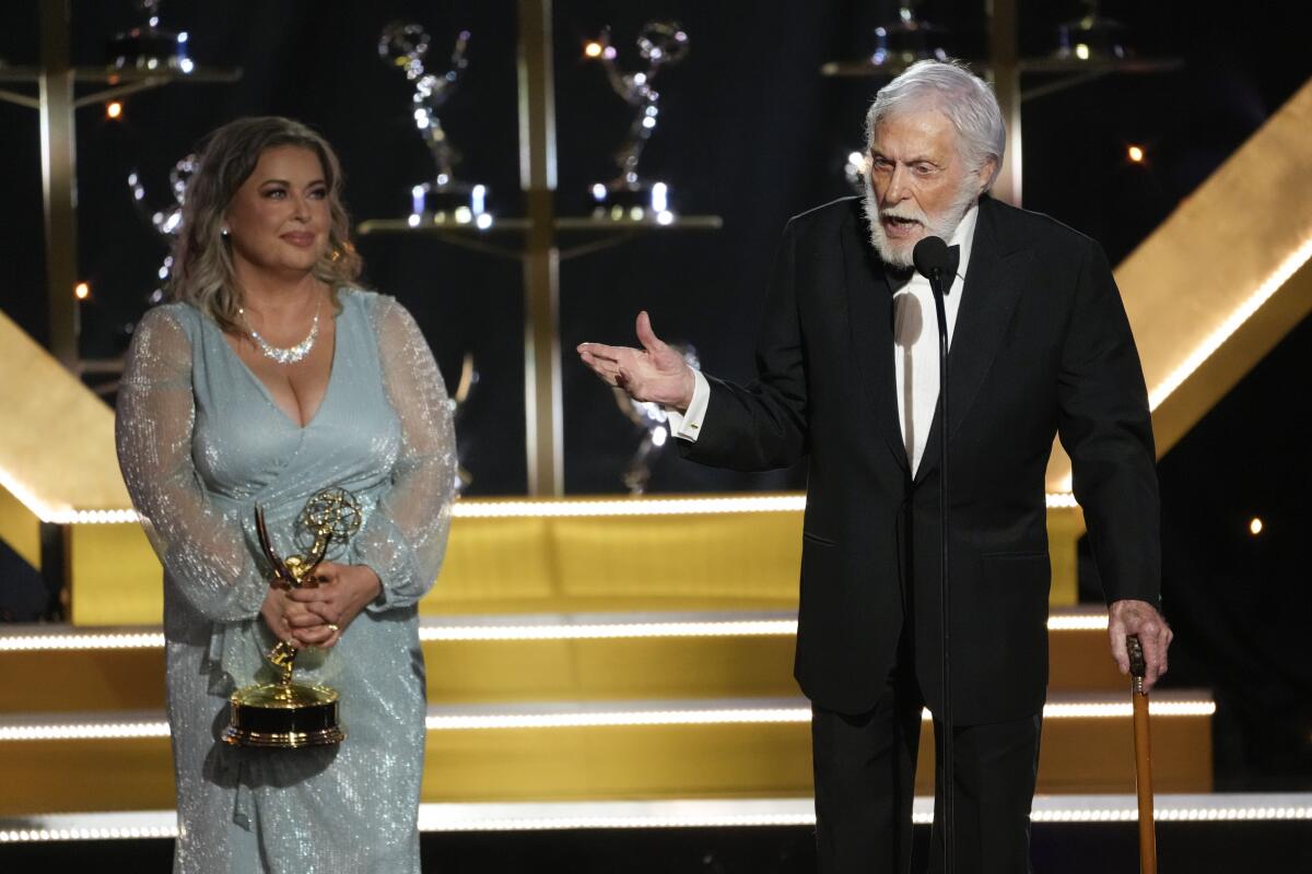Dick Van Dyke speaks into a microphone while wife Arlene Silver stands in a seafoam green dress holding an Emmy statuette
