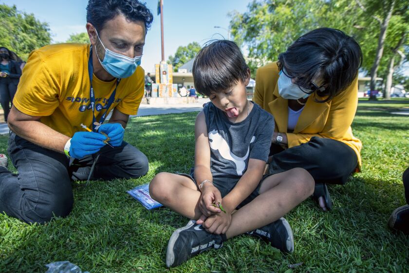 LOS ANGELES, CA - MAY 05: Allan Fernandez, 7, middle, reacts after Dr, David Bolour, left, gives him a vaccination as CDC Director Dr. Rochelle Walensky looks on in Ted Watkins Memorial Park on Thursday, May 5, 2022 in Los Angeles, CA. CDC Director Dr. Rochelle Walensky visted a health department vaccination site in the park. Afterwards she walked into the park and watches children and their parents being vaccinated in the park by an outreach team from the near by site. (Francine Orr / Los Angeles Times)