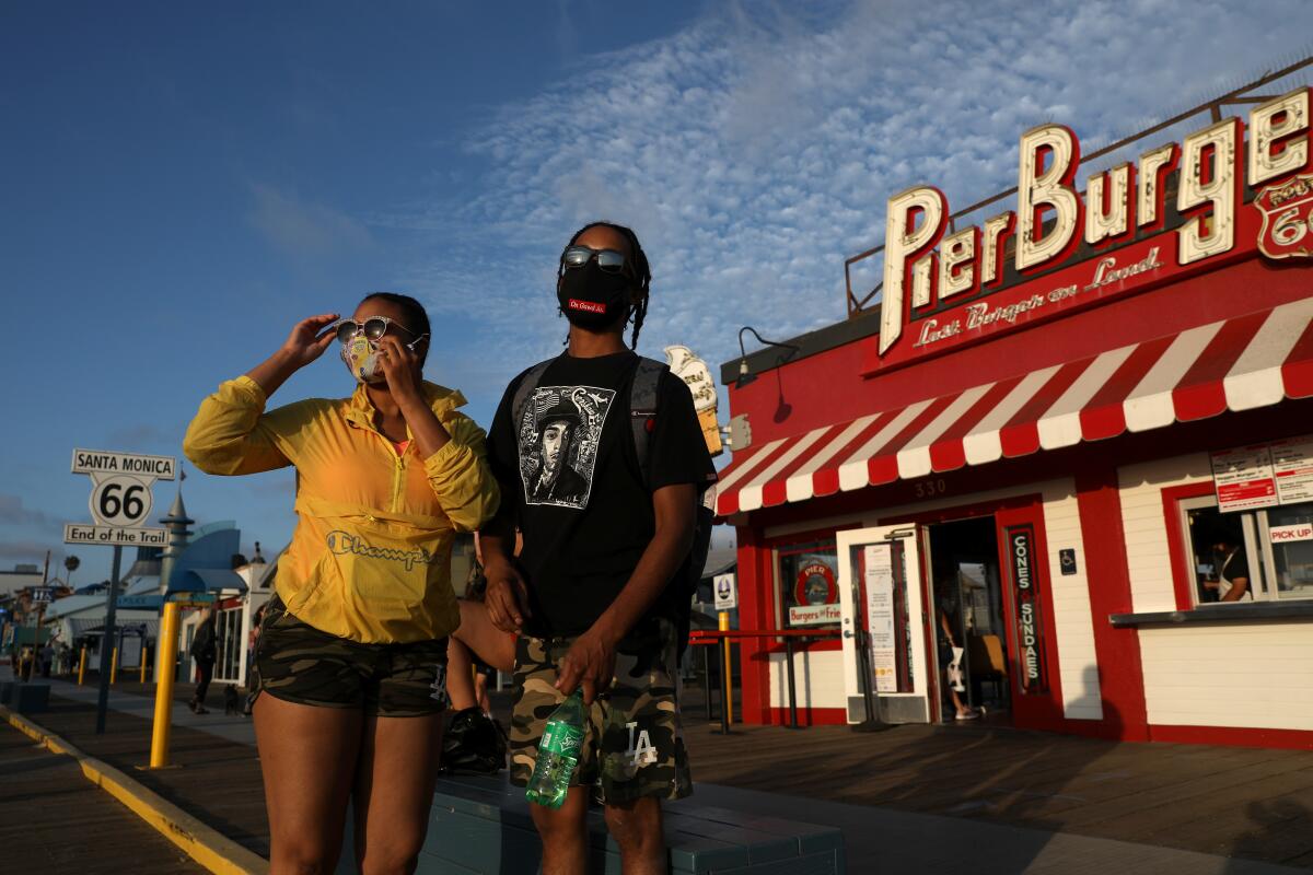 A couple, wearing protective face coverings, stand in front of a row of pierside shops