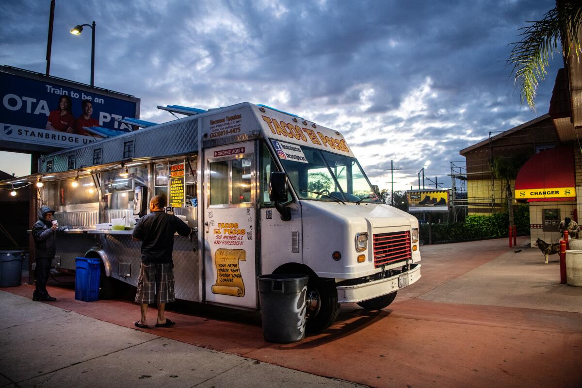 Two people stand in front of a taco truck at dusk.