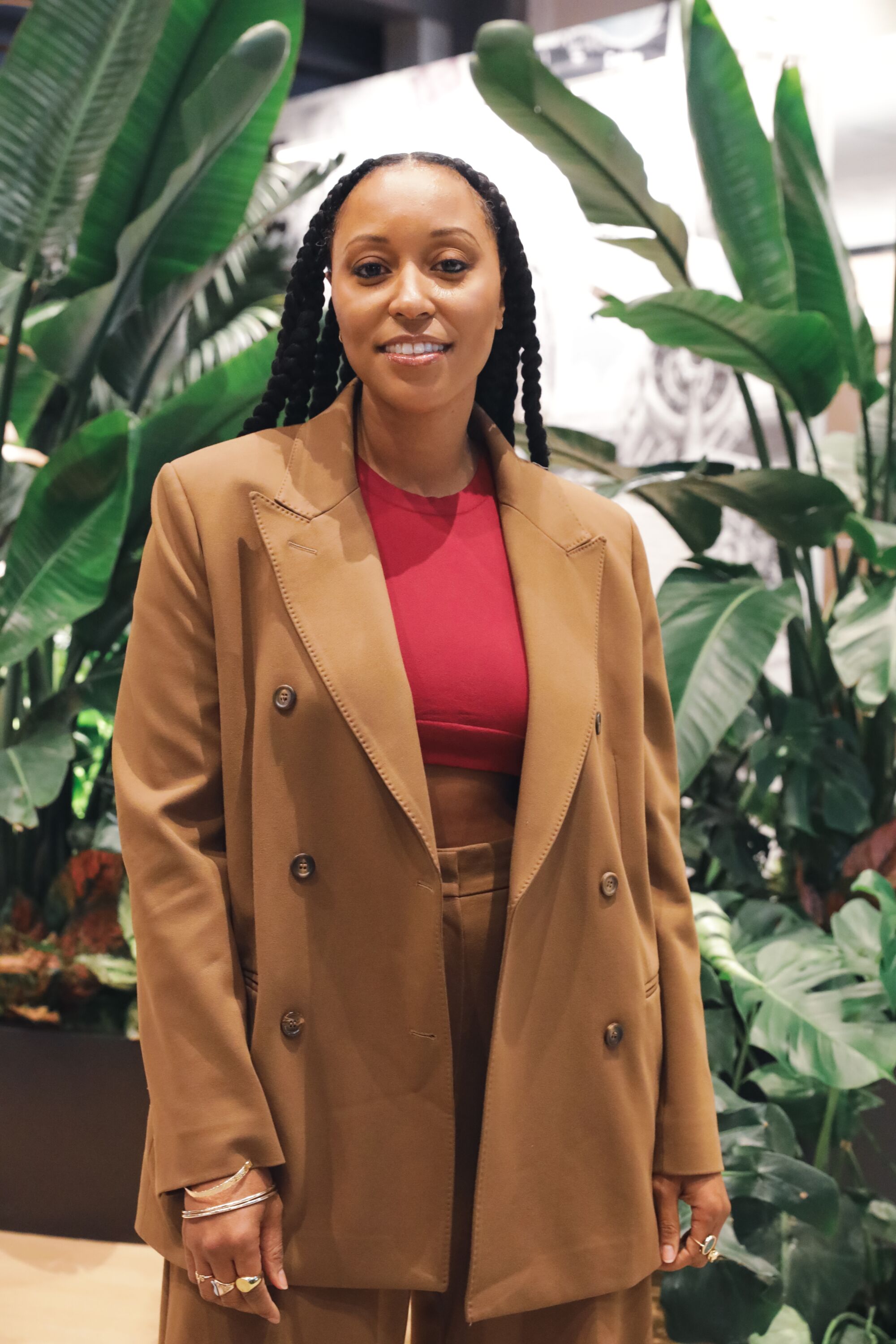 Black woman in a light brown pant suit smiles and poses in front of plants