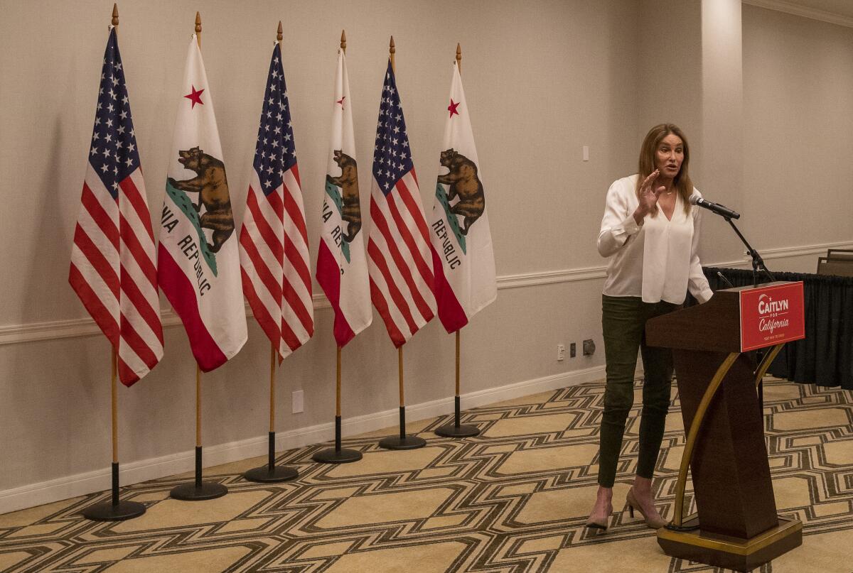 Caitlyn Jenner stands in front of a row of flags at a lectern