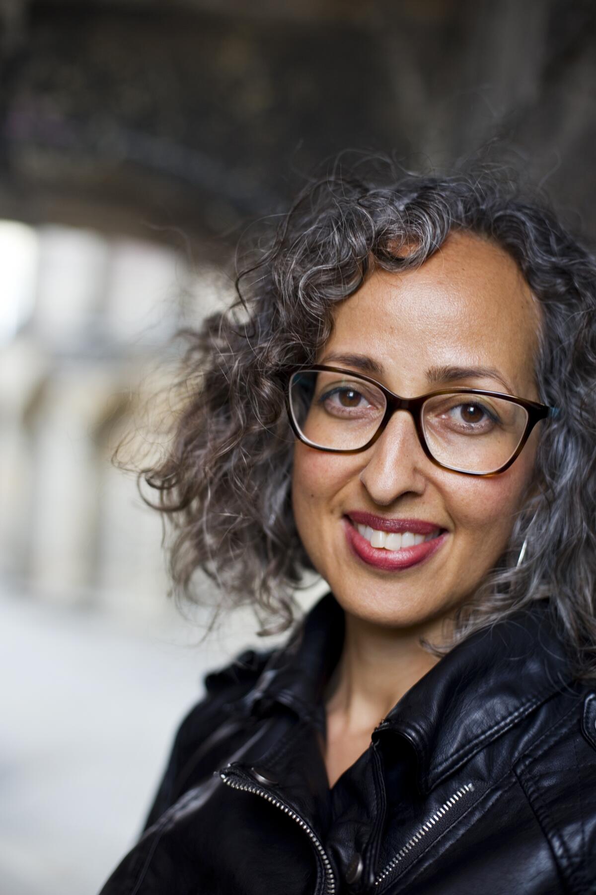 A portrait of a woman with curly hair and glasses and wearing a black jacket.