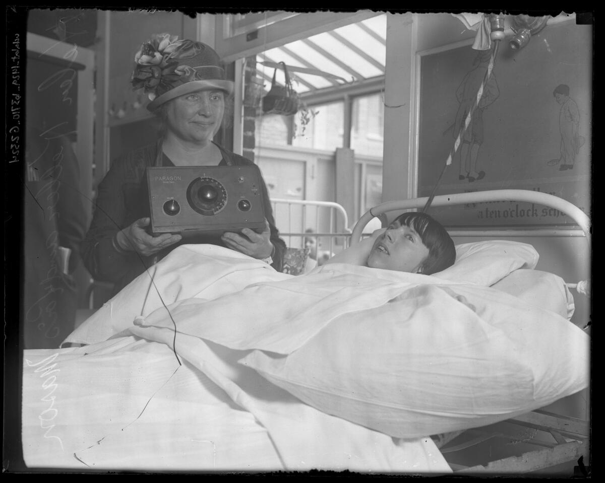 A woman in a hat holds a radio next to a hospital bed where a girl lies covered in white bedding.