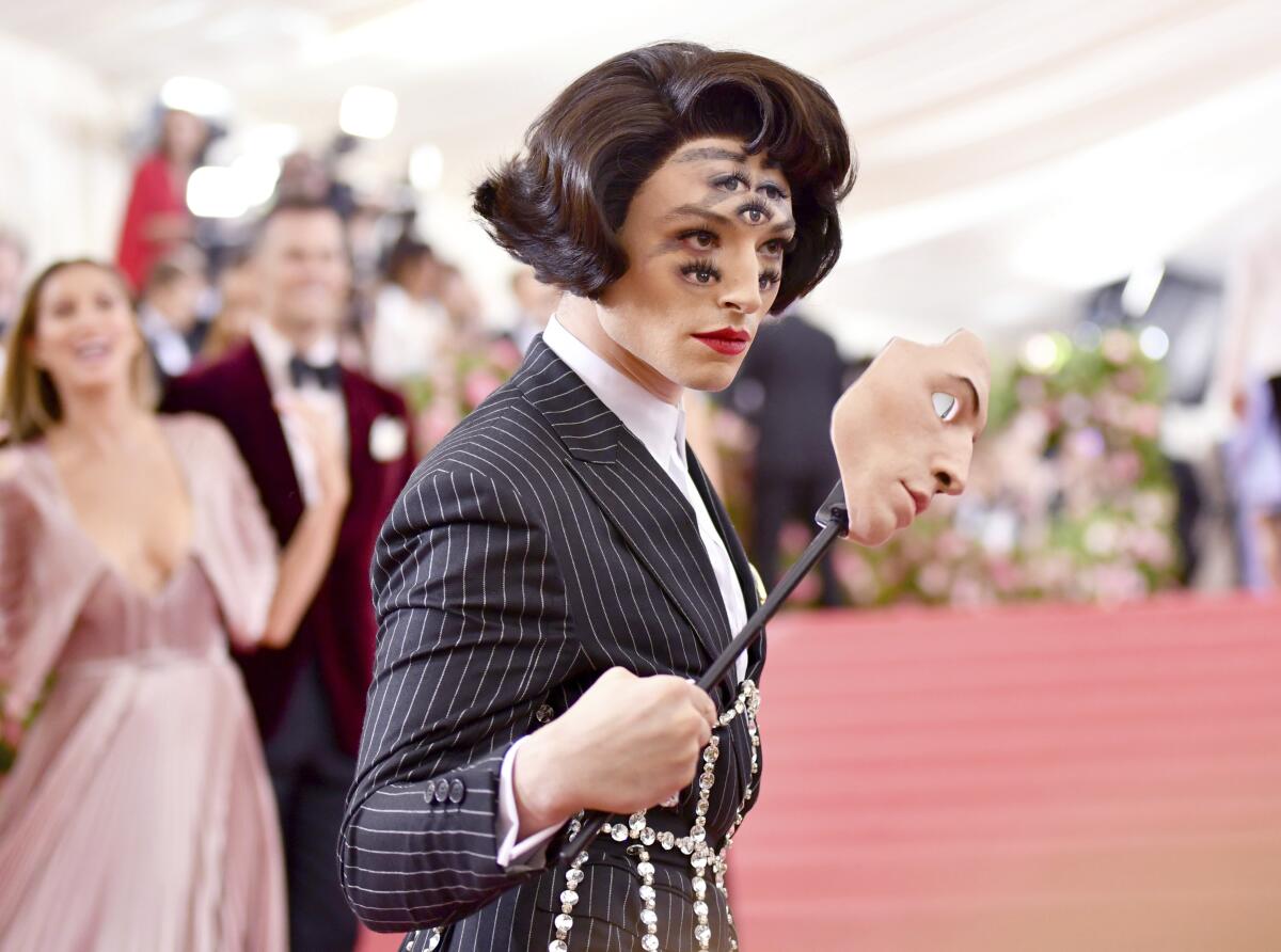 A person in a tuxedo, holding a mask and whose face make-up includes five additional eyes