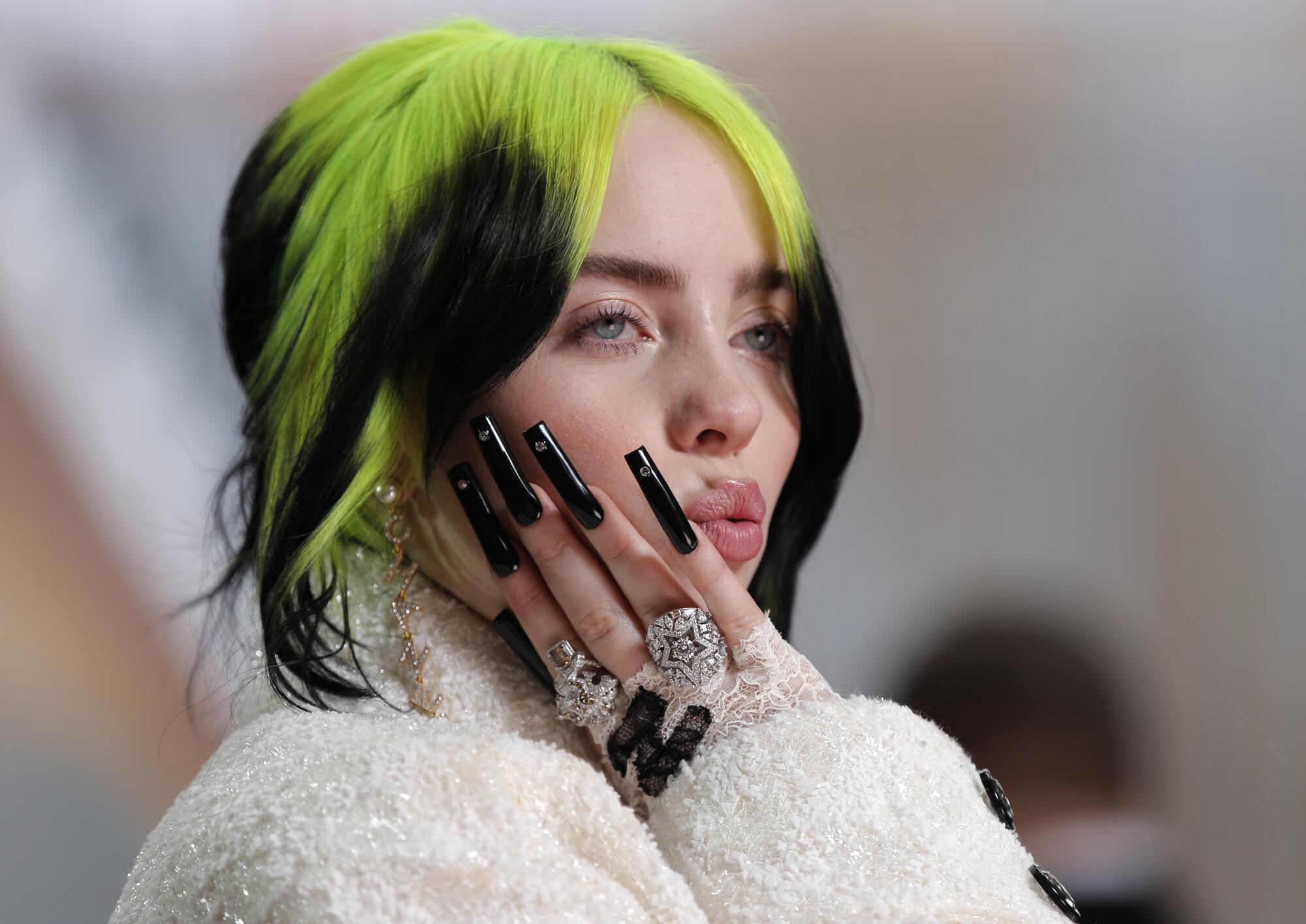 A woman with green and black hair raises a hand with long black nails to her face.