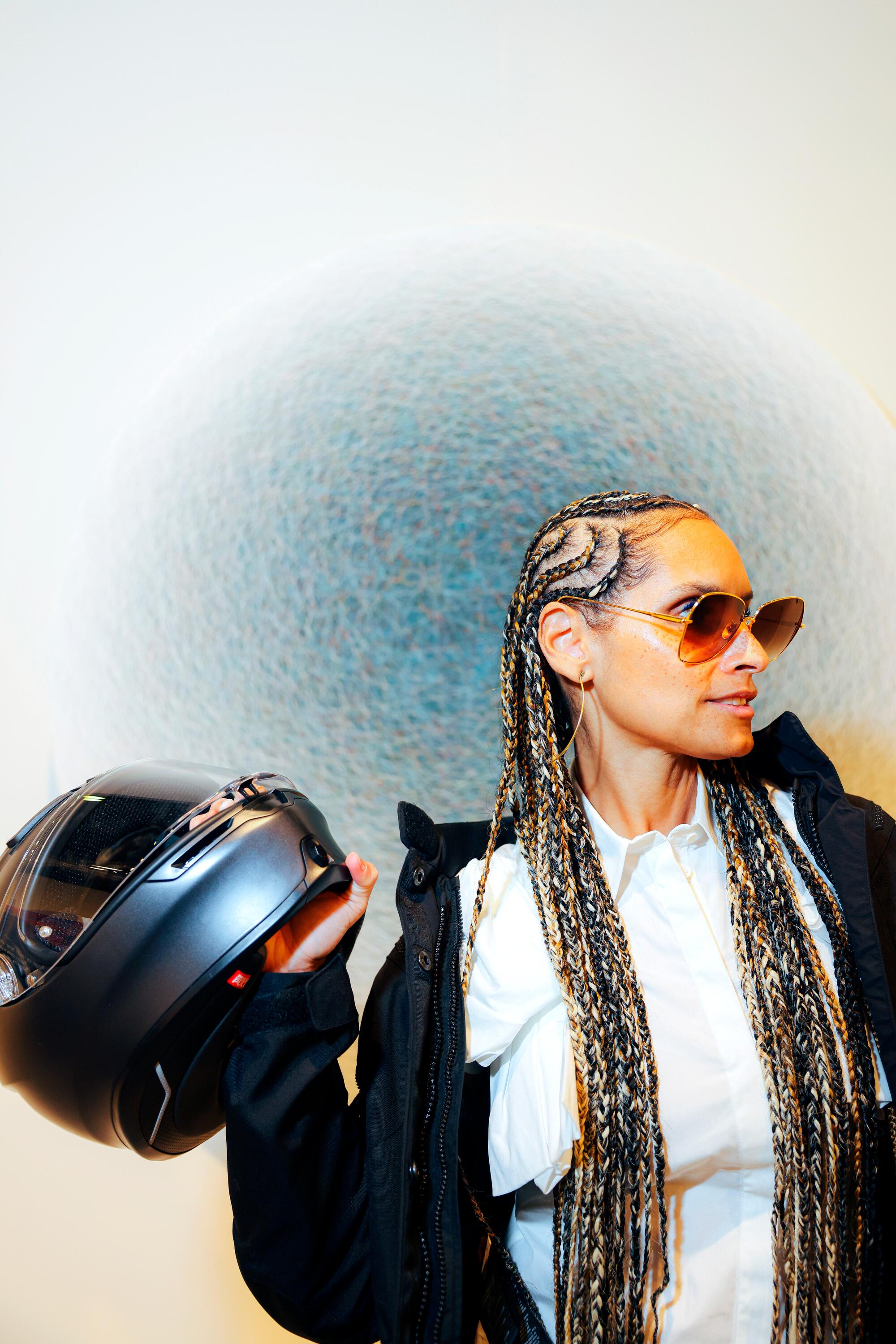A person with long braids poses in front of a work of art holding a motorcycle helmet.