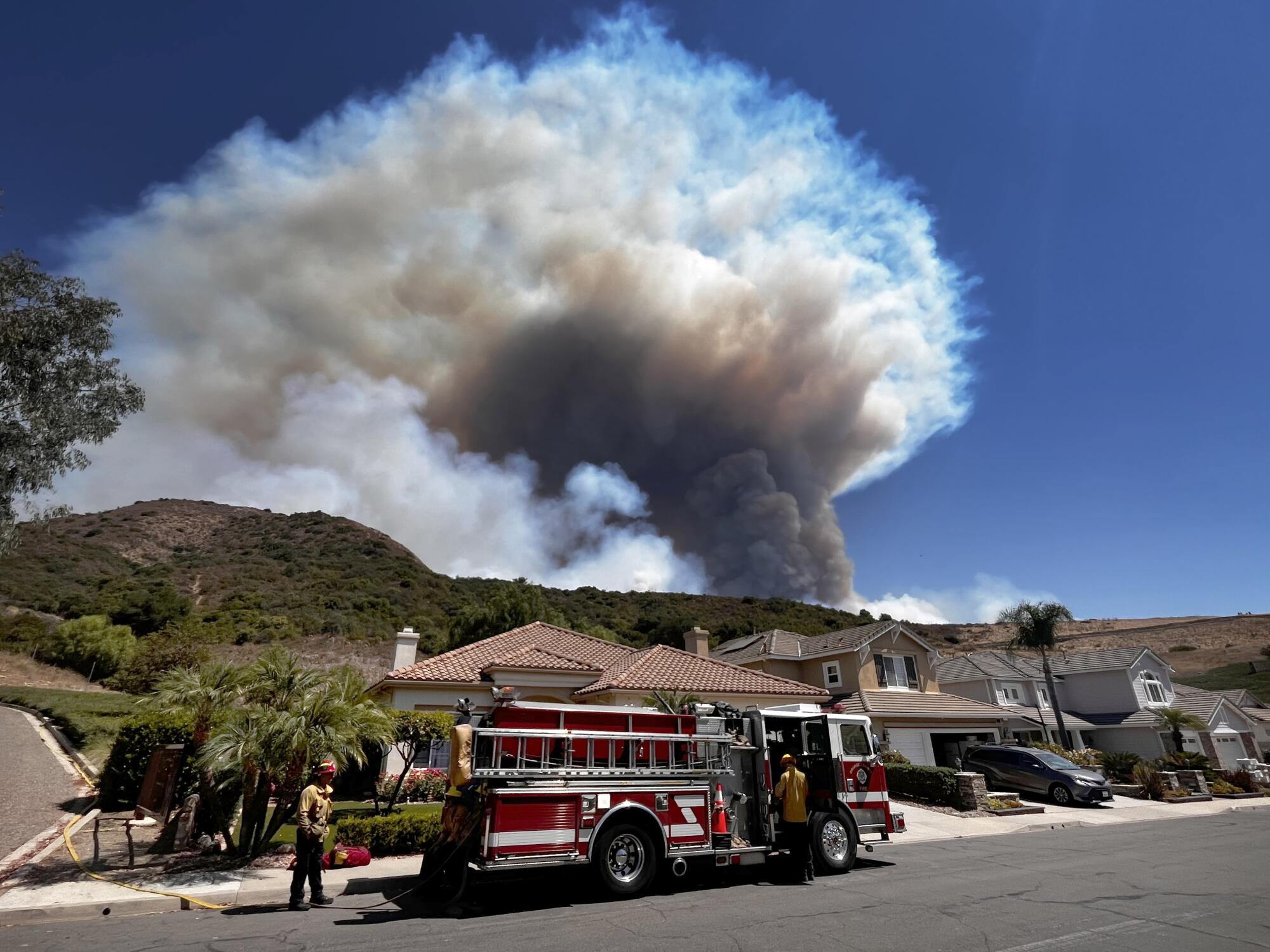 A fire truck on a residential street with a huge smoke plume in the distance.