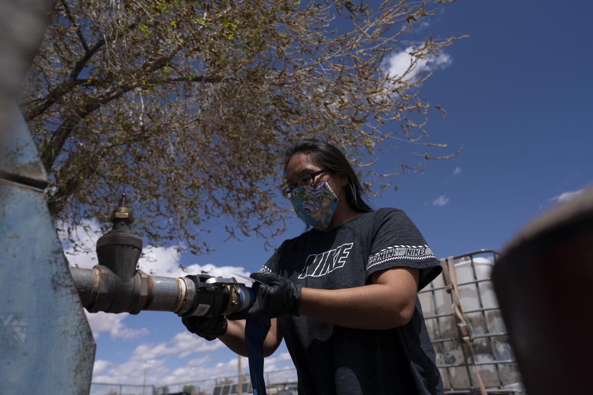 A woman in mask and T-shirt stands outdoors beside a water tank.