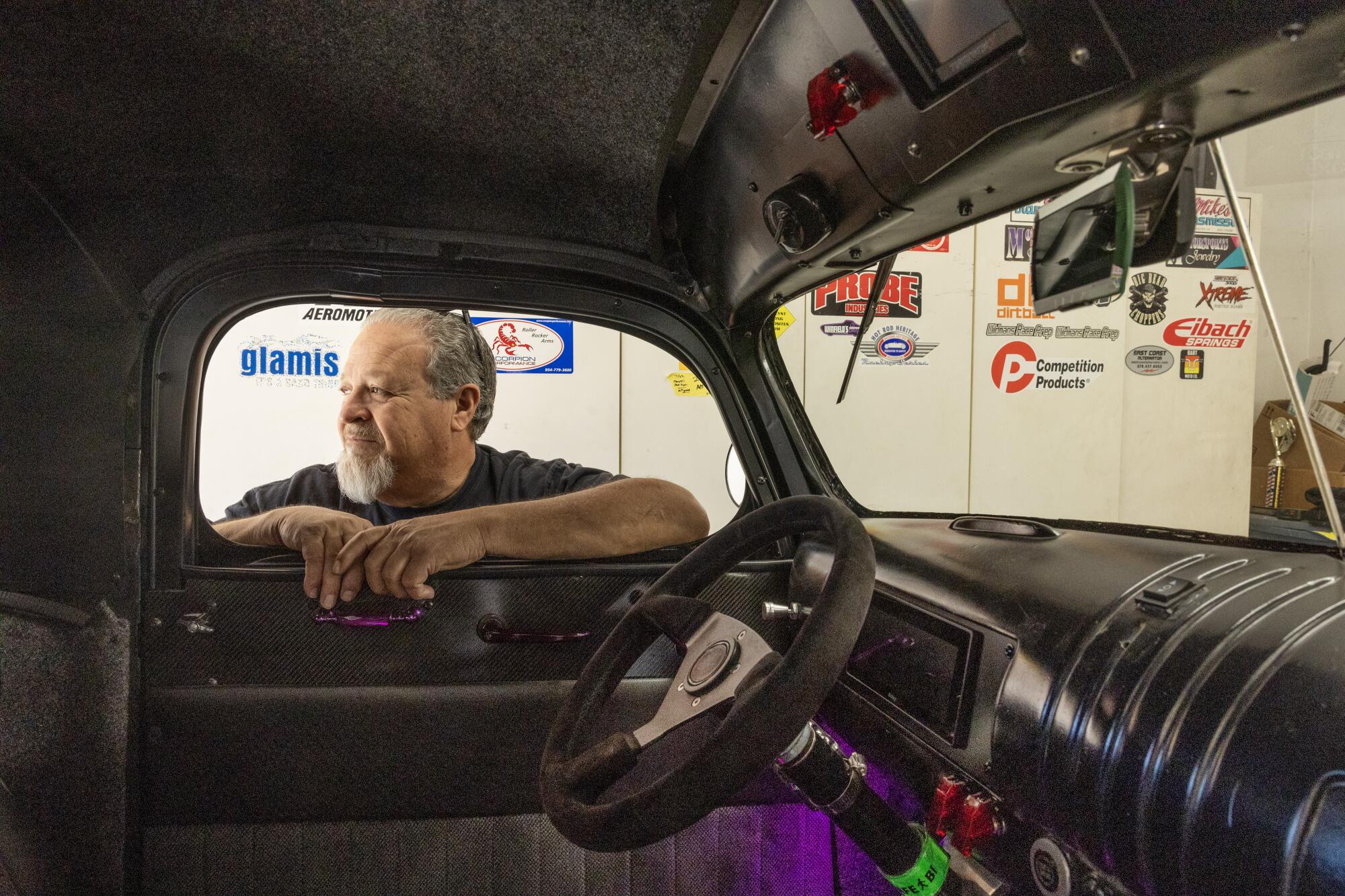 A man with gray hair and beard standing outside his vehicle leans on the window on the driver's side