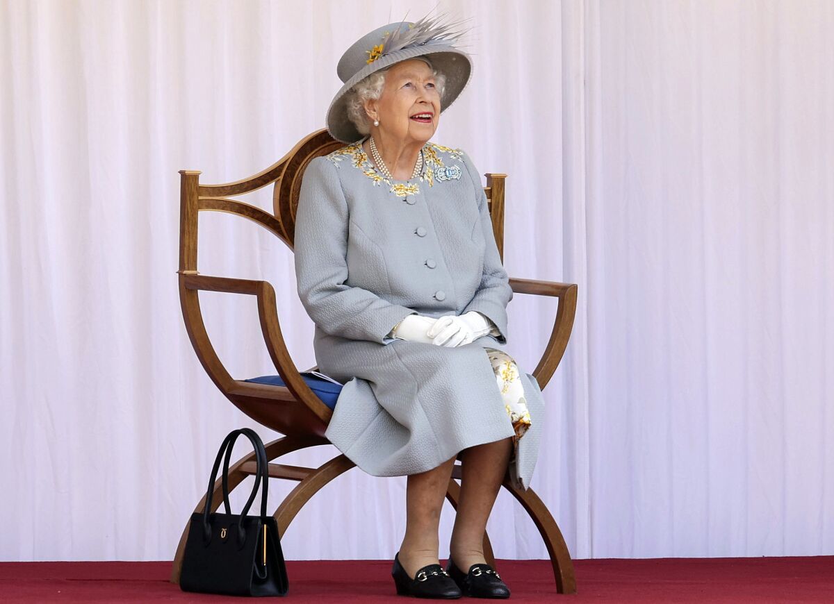 An elderly woman, seated, wearing a hat, looking up,  gloved hands clasped on her lap, with a handbag by the chair.  