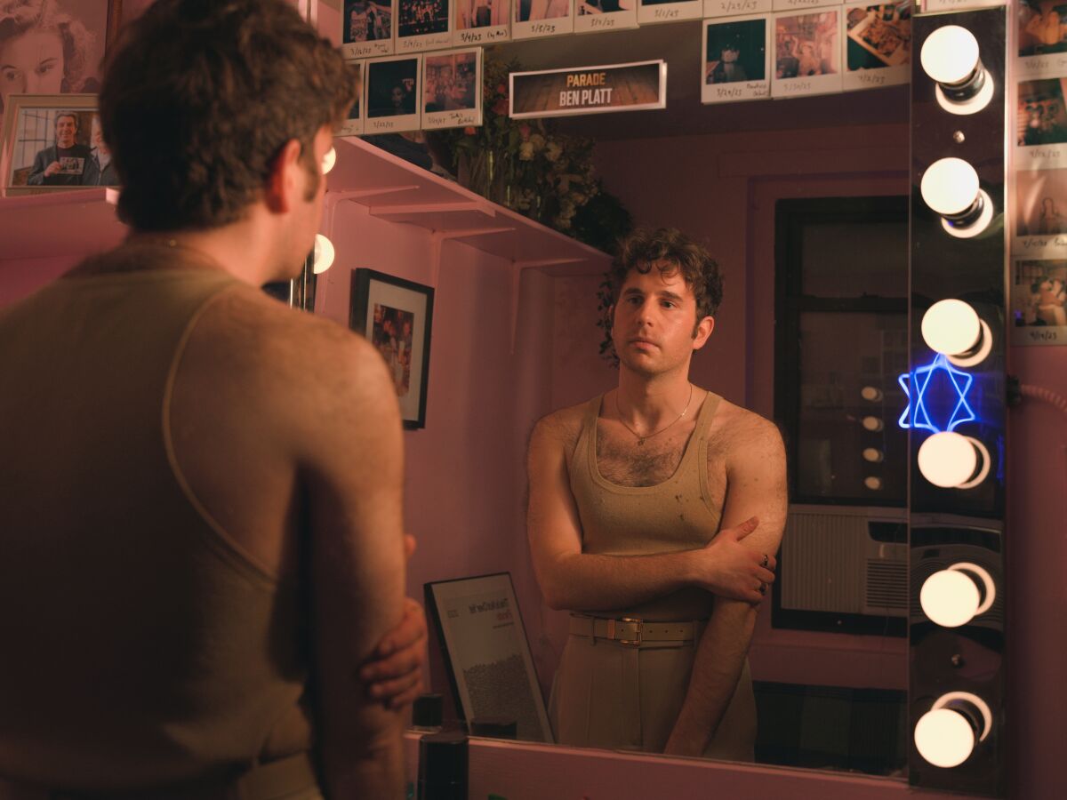 A man in beige tank top looks into the mirror, a neon Star of David illuminated behind him