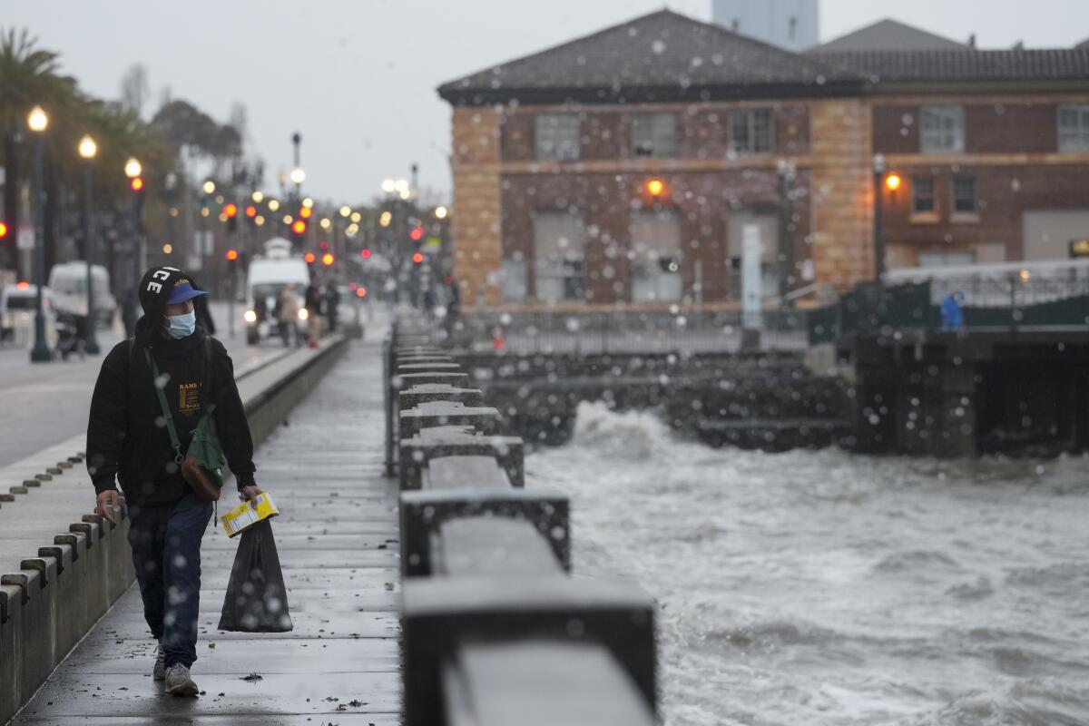 A person in a heavy hooded jacket is seen on a city walkway that borders turbulent water. 