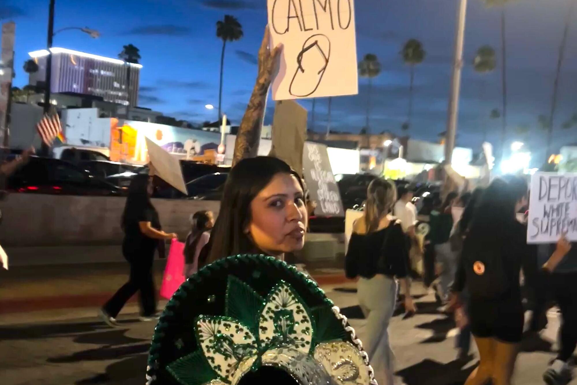 A woman stands at a protest with a person holding a sign written in Spanish.