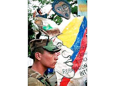 A Colombian soldier observes a ceremony commemorating the third anniversary of the deaths of 18 people in Santo Domingo. In back, a boy with his face painted white hangs a sign protesting the attack.
