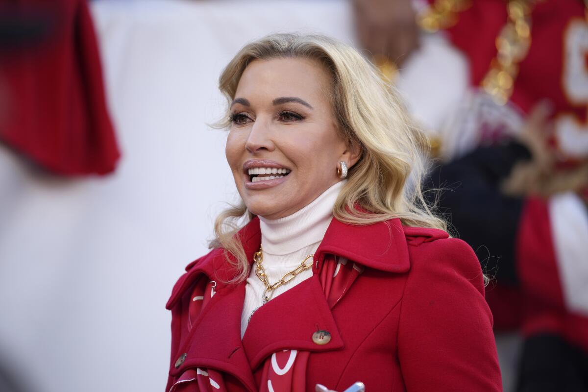 Uma mulher loira vestindo uma gola alta branca e uma jaqueta vermelha está sorrindo