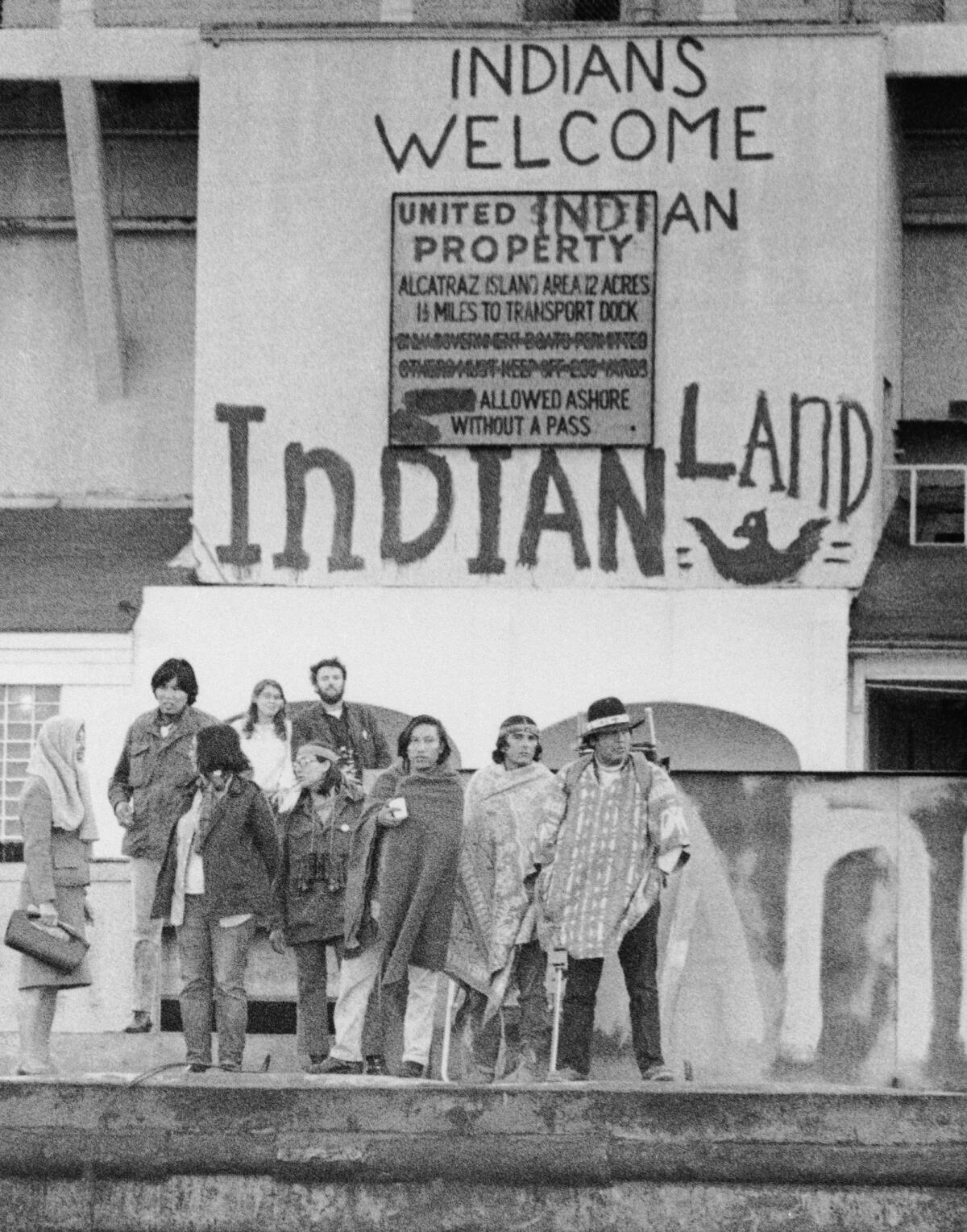 A group of Indigenous people stand under a sign that reads "INDIANS WELCOME" on the docks at Alcatraz