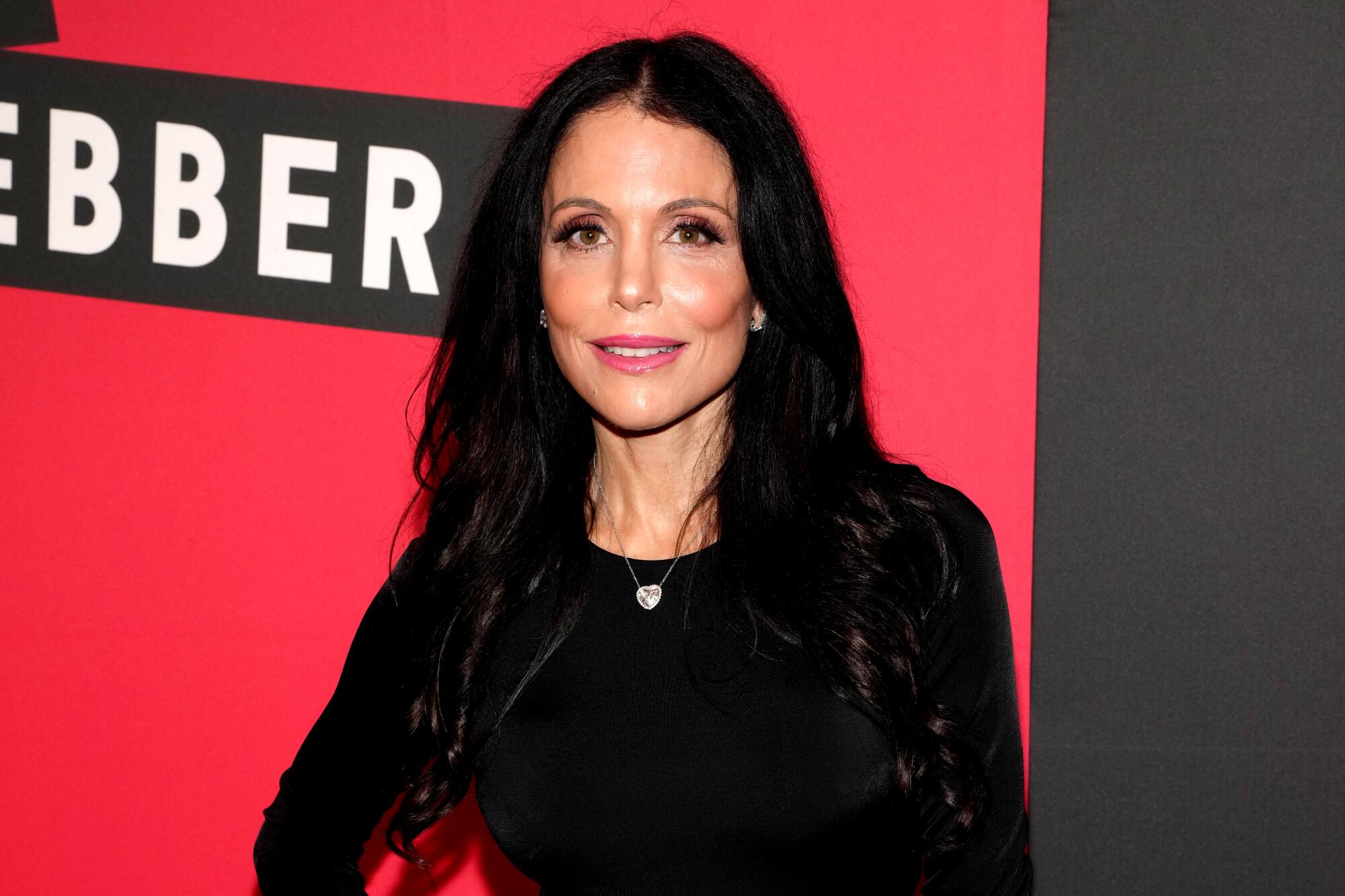 Bethenny Frankel in a black top and silver necklace stands against a red backdrop. 