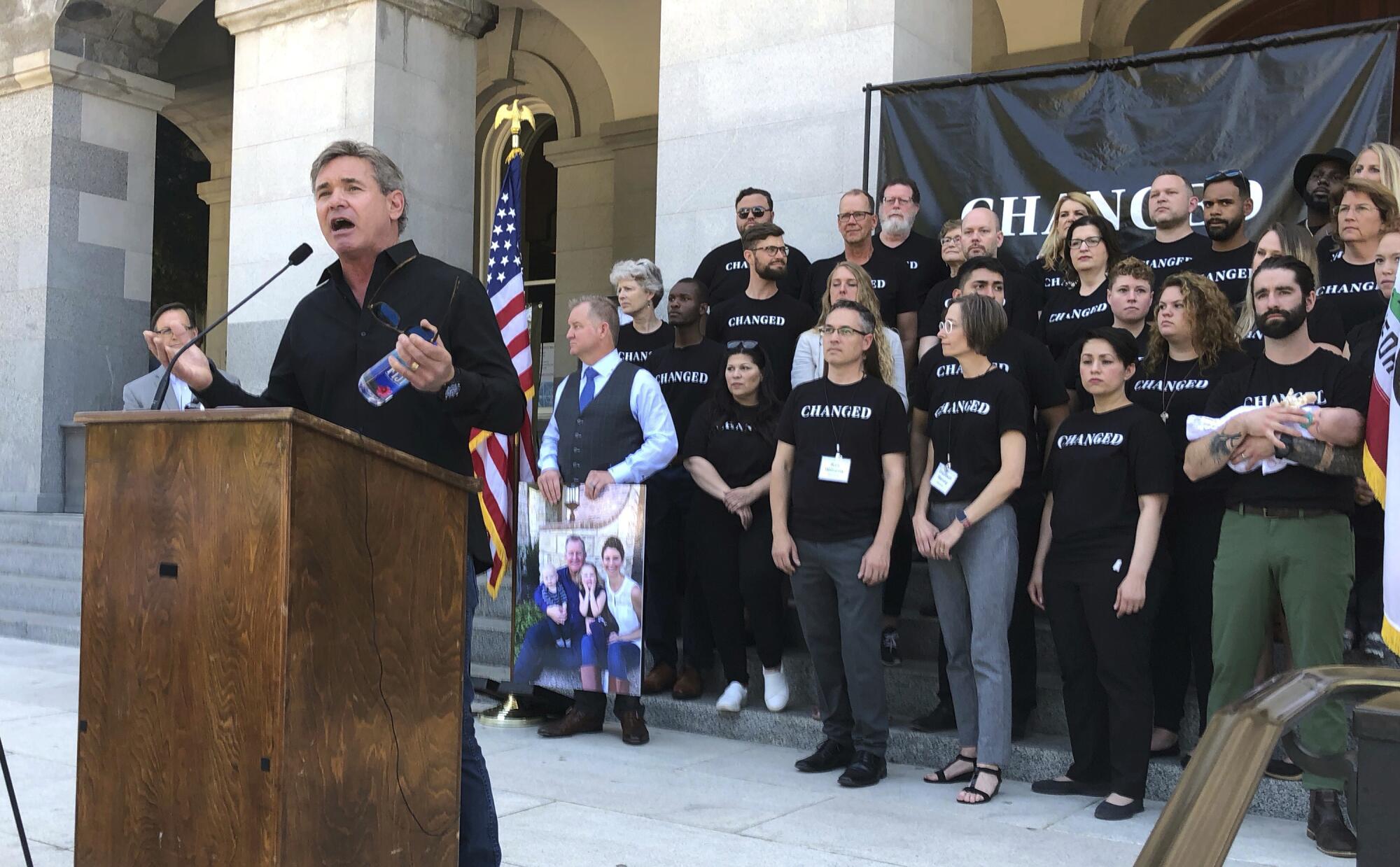 A man speaks at a lectern while people dressed in matching shirts stand behind him.