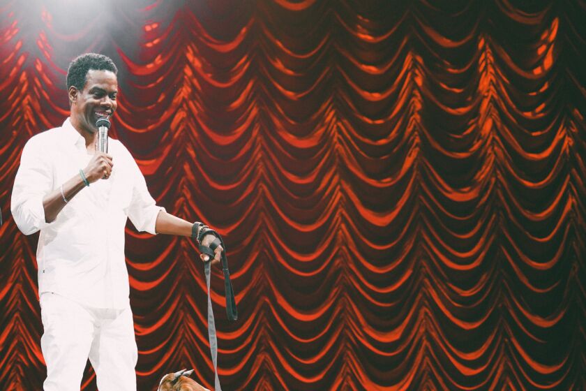 A Black man in a white outfit holds a microphone in one hand and a goat on a leash in the other