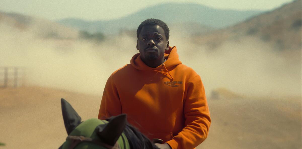A man in an orange hoodie riding a horse in the desert.
