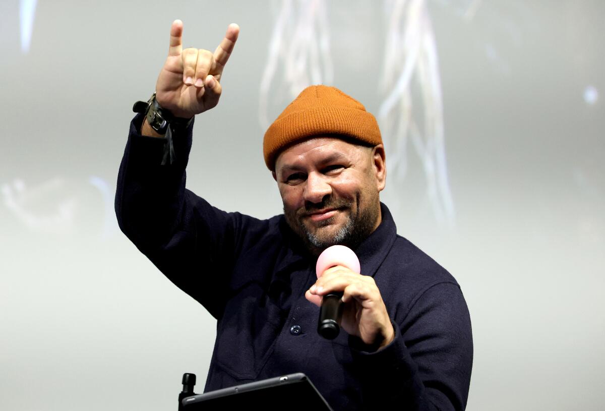 A man in an orange beanie and dark outfit holding a microphone with his left hand and putting up the rock sign with his right