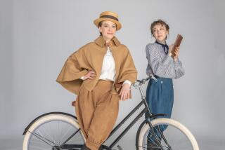 Two actresses in 1890s-style costumes pose with a bicycle before a gray backdrop.