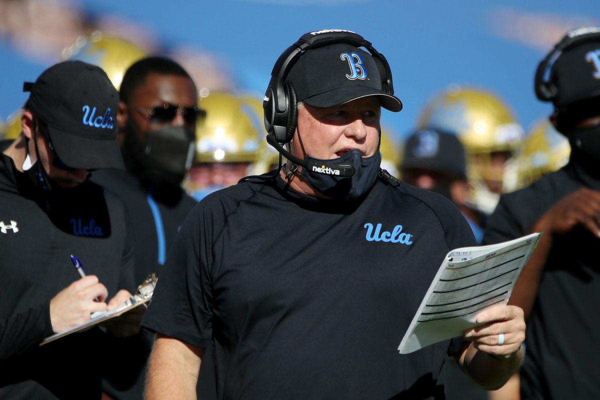 UCLA football coach Chip Kelly reads a play from a chart while standing on the sideline.