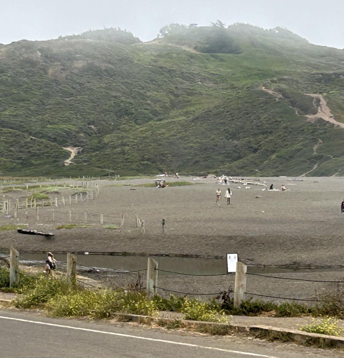 People walk and sit on a beach below a green hill with low fog overhead.